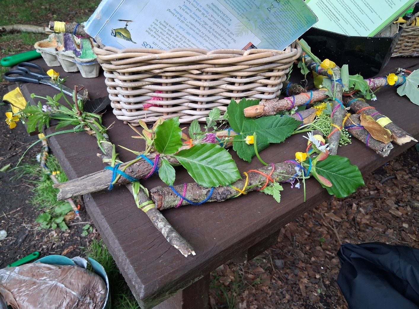 Nature-craft creations on a wooden table: sticks wrapped with colourful bands, leaves and wildflowers, beside a woven basket and nature journals.