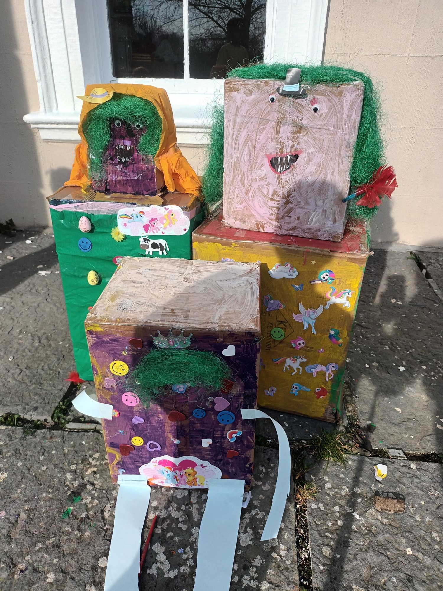 The completed cardboard family of decorated boxes with woolly hair and stickers, displayed outdoors in the sunshine.