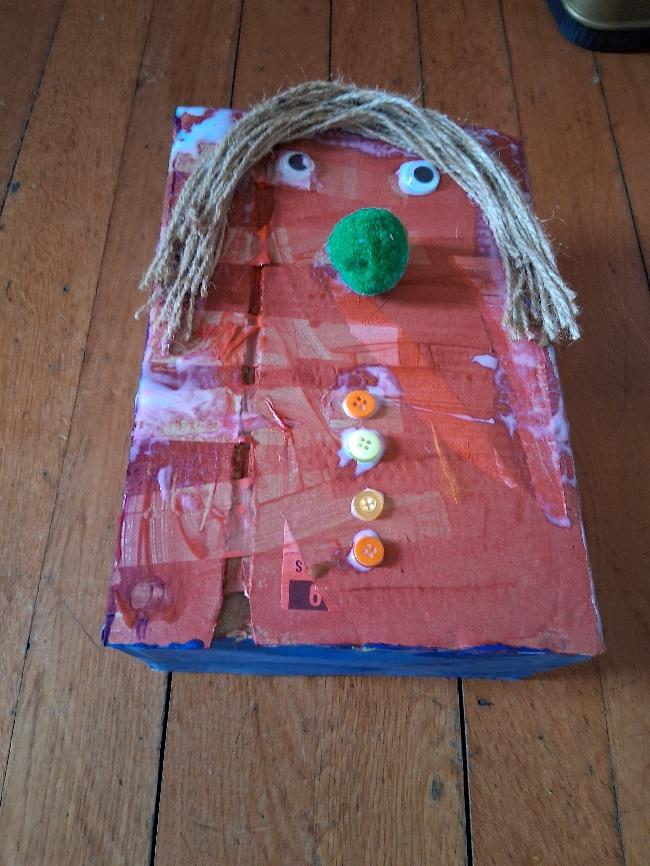 A small cardboard box decorated as a head, with twine for hair, googly eyes and coloured buttons.