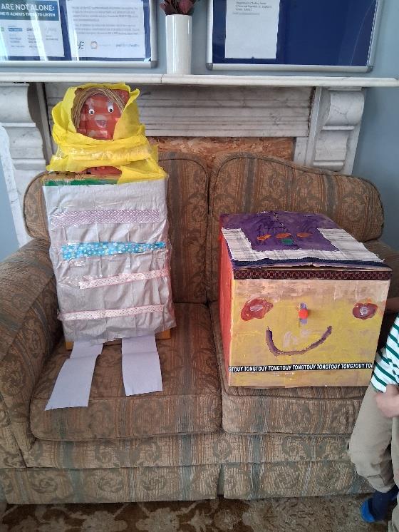 The two finished cardboard figures, the husband and wife, dressed in fabric and paper and standing on a sofa.