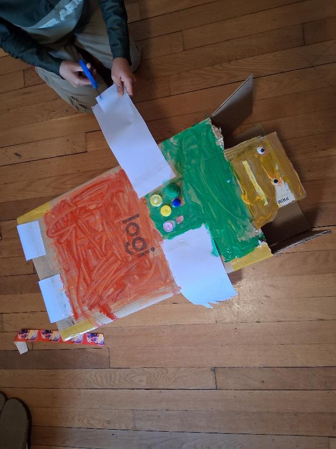 A child sitting on the floor beside the cardboard robot they built and painted in orange and green, with button features and a paper face.