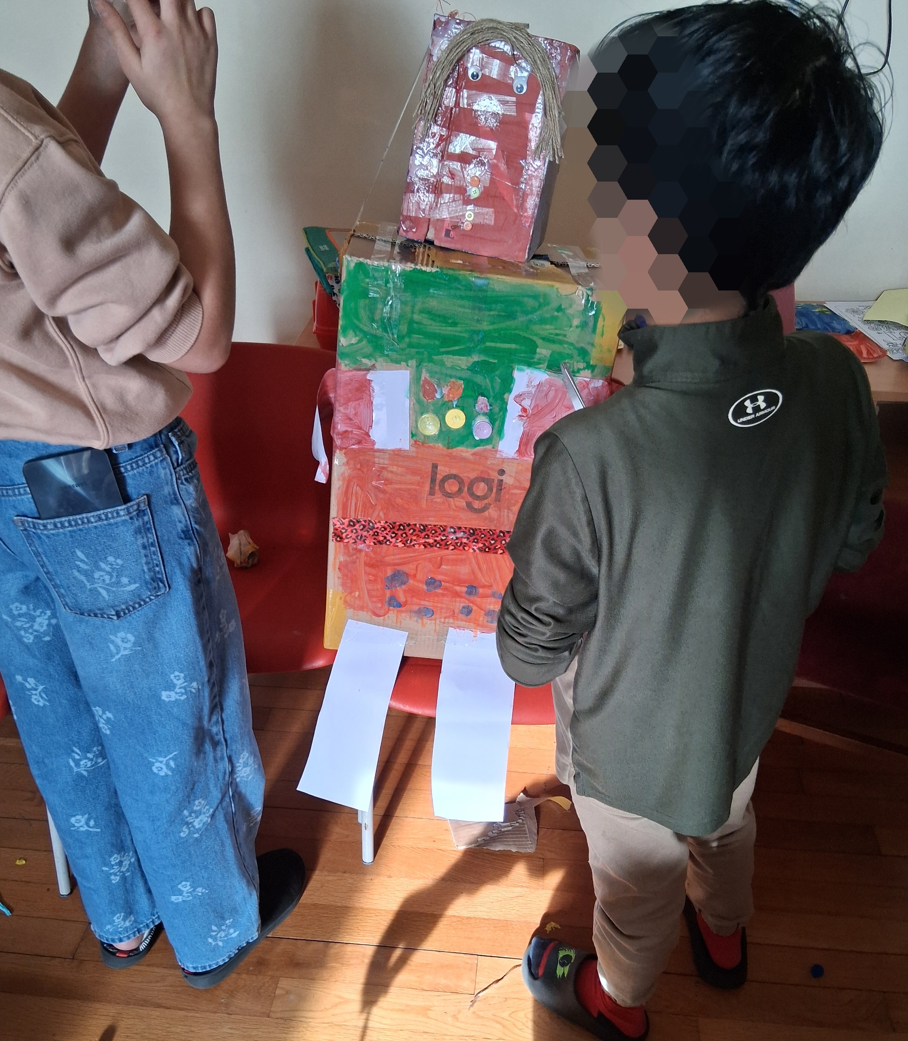 Two children standing either side of the tall cardboard robot figure they have dressed and decorated together.
